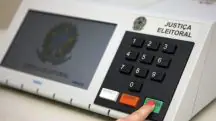 Modelo de urna eletrônica usada no sistema eleitoral brasileiro. Na imagem, mostra-se um dedo indicador direito, possivelmente de uma mulher, pois está com esmalte vermelho na unha, em que simula-se o momento final da votação: teclando o botão verde, de 'confirma'