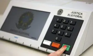 Modelo de urna eletrônica usada no sistema eleitoral brasileiro. Na imagem, mostra-se um dedo indicador direito, possivelmente de uma mulher, pois está com esmalte vermelho na unha, em que simula-se o momento final da votação: teclando o botão verde, de 'confirma'