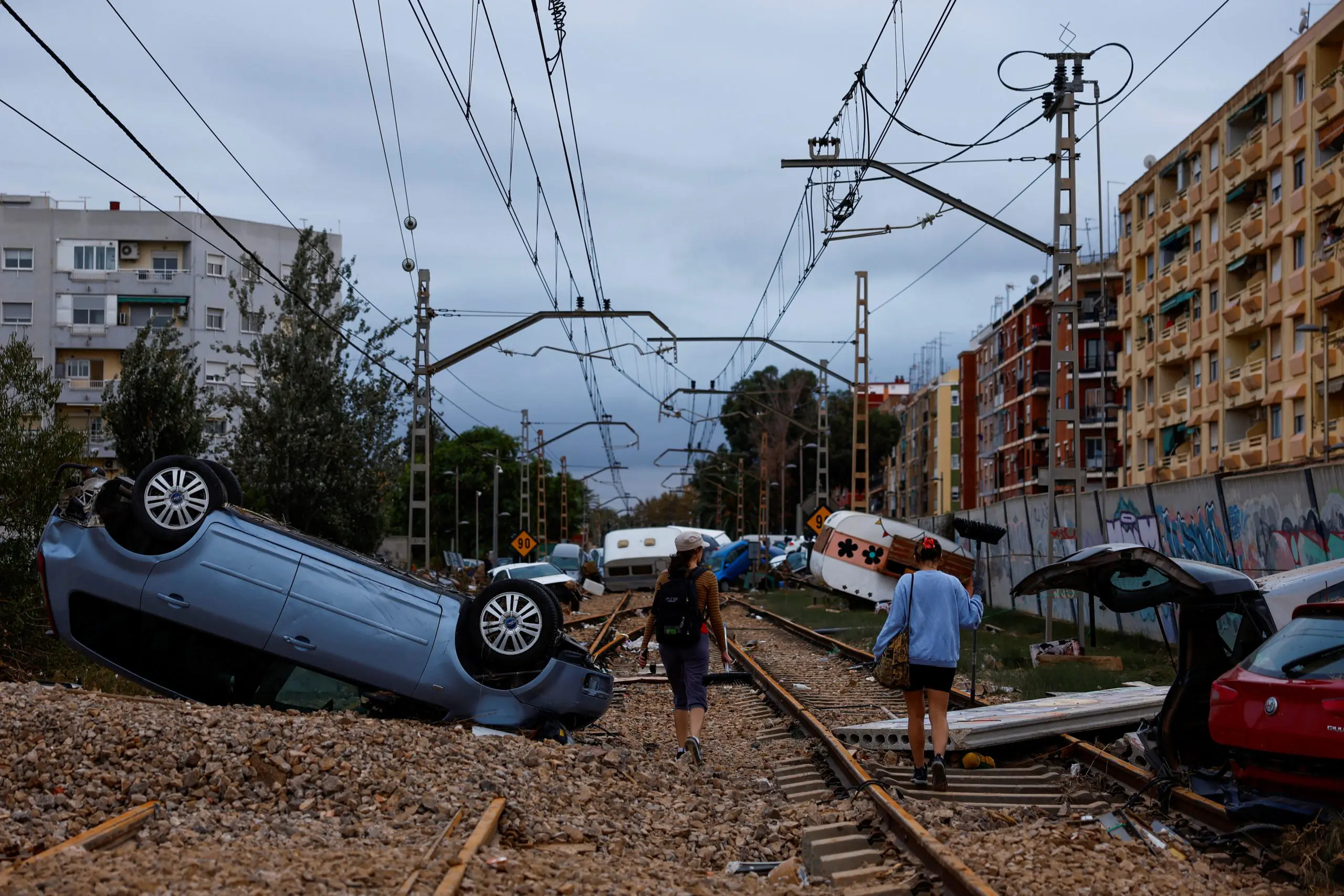 Mulheres com vassouras passam por carros encalhados ao longo da ferrovia, em Alfafar, Espanha - 31/10/2024 | Foto: Susana Vera/Reuters