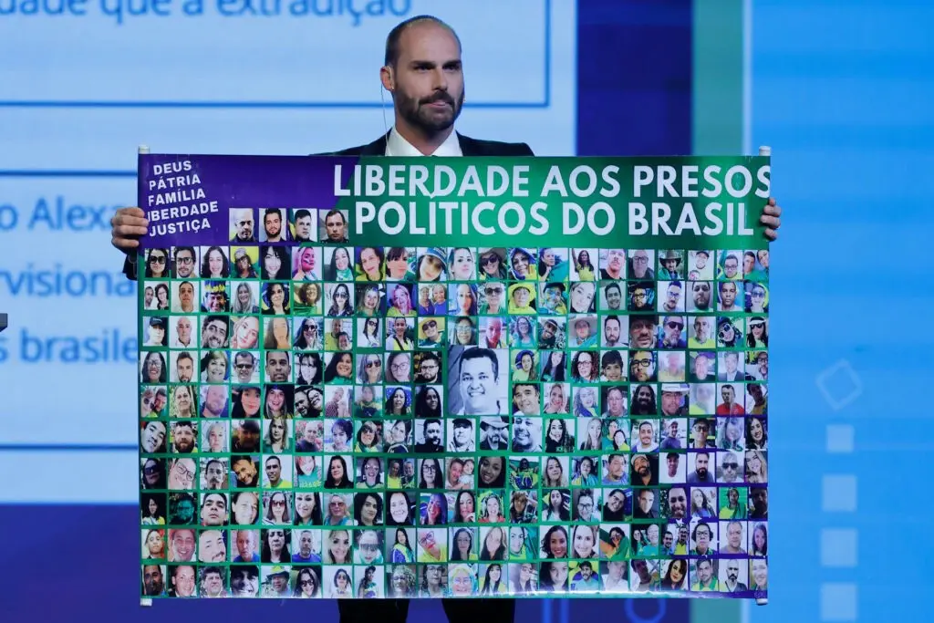 Durante conferência conservadora em Buenos Aires, na Argentina, Eduardo Bolsonaro (PL) mostra cartaz com imagens dos manifestantes que estaria sendo tratados como terroristas pelo STF | Foto: Reprodução/Redes sociais