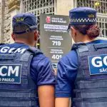 Dois guardas civis municipais de costas para a câmera e olhando para o "prisômetro", painel criado pela Prefeitura de São Paulo, para exibir número de pessoas presas com o auxílio do programa Smart Sampa
