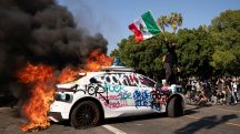 Um manifestante agita uma bandeira mexicana no topo de um veículo em chamas durante um protesto no centro de Los Angeles, Califórnia - 8/6/2025 | Foto: David Ryder/Reuters
