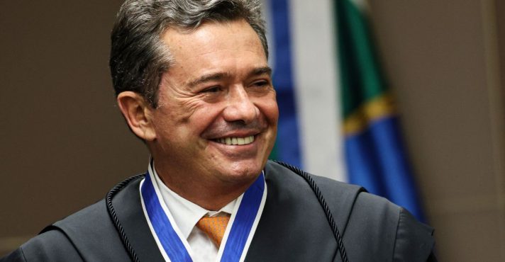 Magistrado brasileiro sorrindo durante cerimônia oficial, usando toga preta e medalha institucional no pescoço