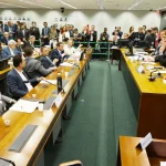 Reunião de comissão na Câmara dos Deputados com parlamentares sentados à mesa, debate em andamento e presença da imprensa.