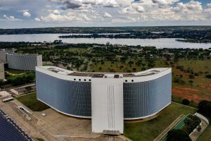 Vista aérea da sede do Tribunal Superior Eleitoral em Brasília, com edifício curvo, área verde ao redor e lago ao fundo.