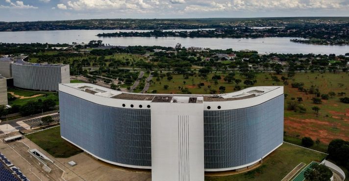Vista aérea da sede do Tribunal Superior Eleitoral em Brasília, com edifício curvo, área verde ao redor e lago ao fundo.