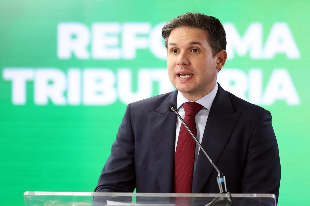 Homem de terno discursando em púlpito com microfone durante pronunciamento oficial, com painel verde institucional ao fundo.