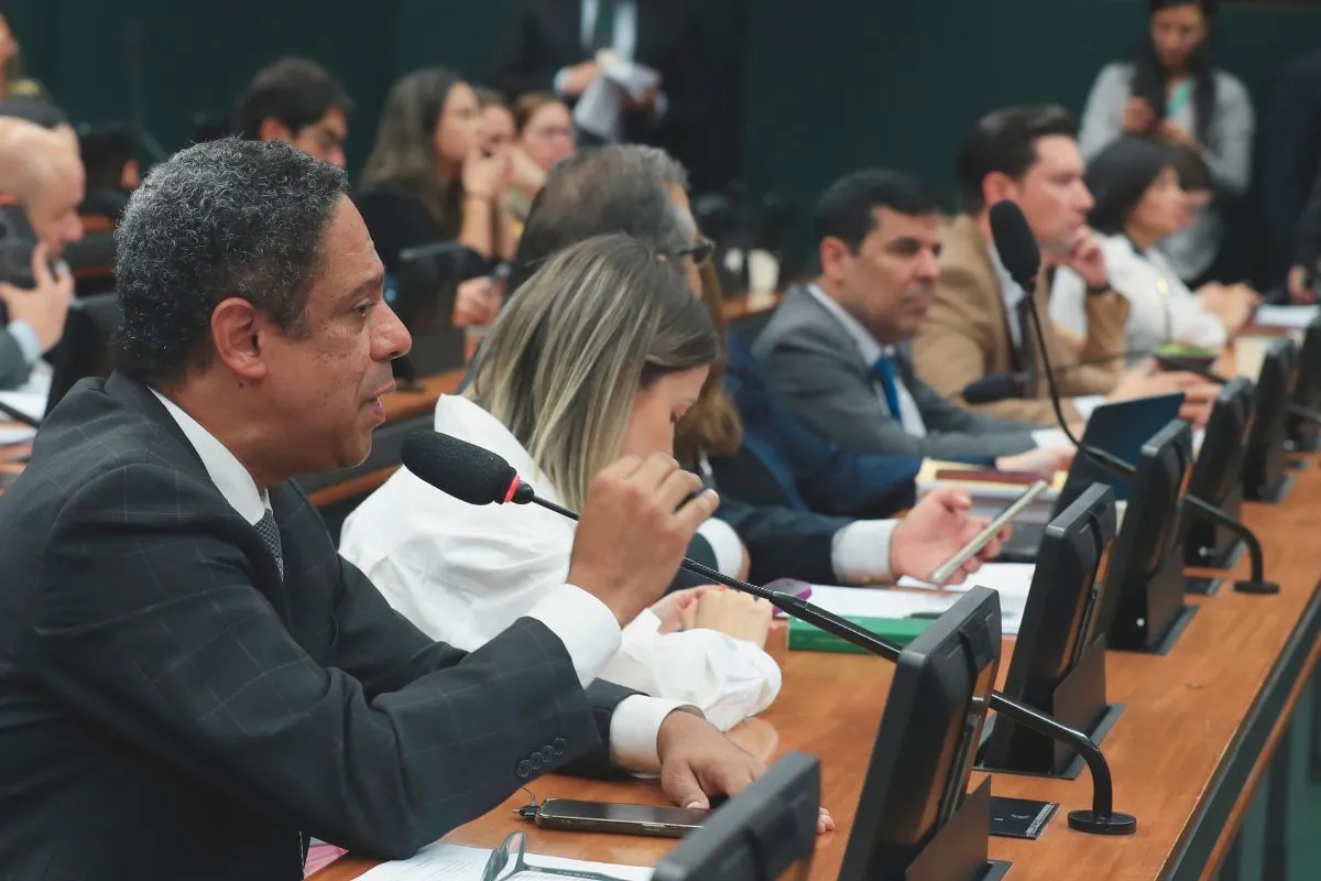 Parlamentar fala ao microfone durante reunião de comissão da Câmara, enquanto outros participantes acompanham a sessão com documentos e monitores.