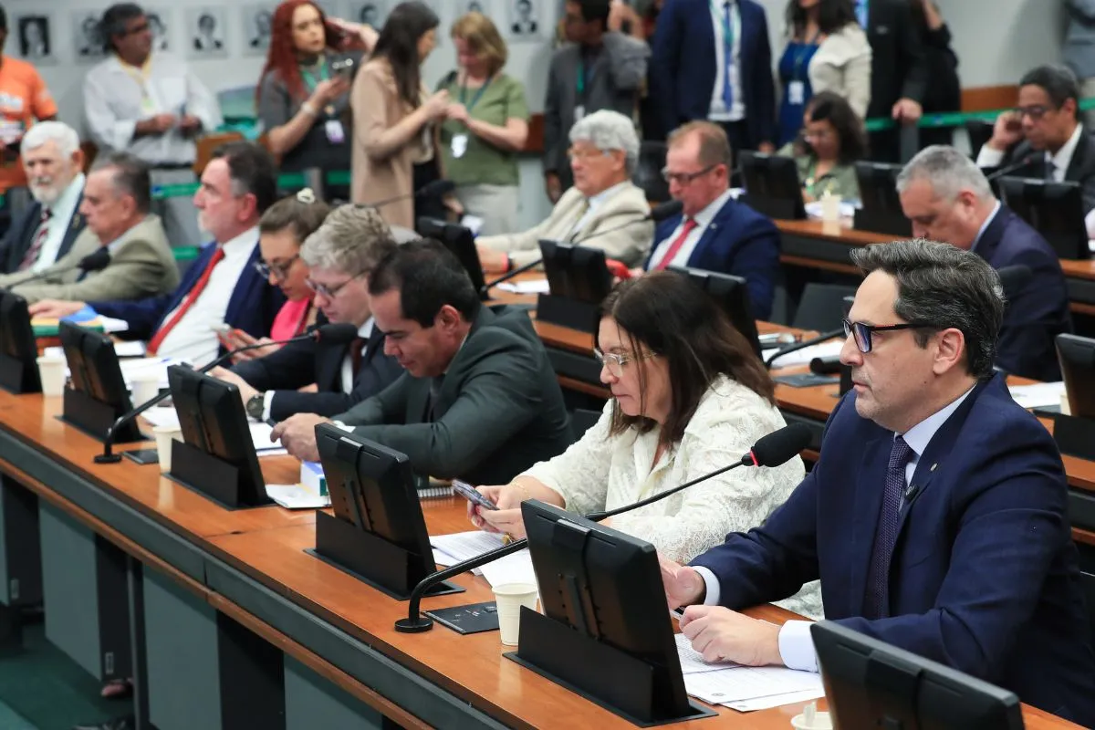 Parlamentares sentados em comissão da Câmara dos Deputados analisam documentos e utilizam monitores durante reunião legislativa.