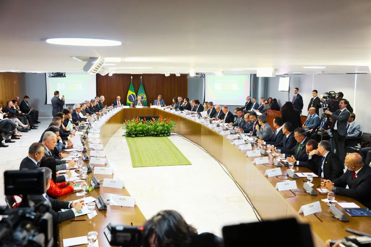 Reunião do governo brasileiro com ministros e autoridades em sala oficial, bandeiras do Brasil ao fundo