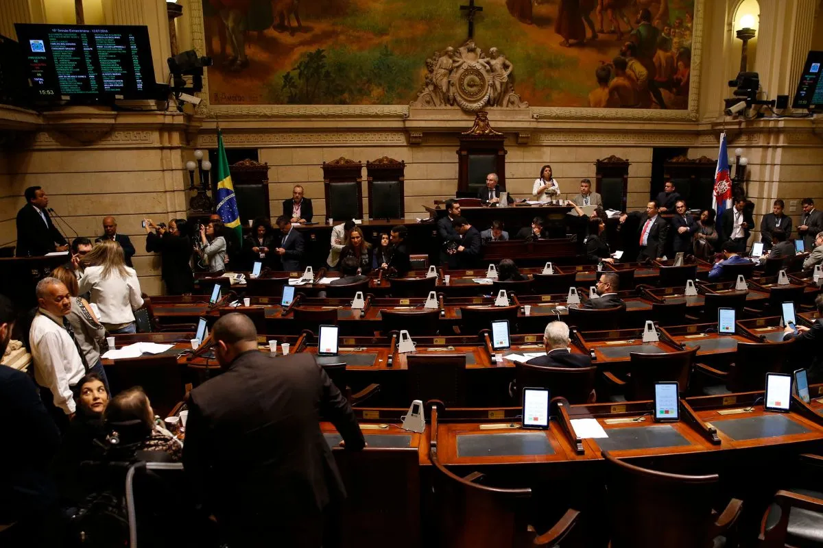 Plenário da Câmara dos Deputados durante sessão legislativa, com parlamentares, painéis eletrônicos, bandeira do Brasil e mesas com tablets.