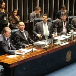 Parlamentares participam de sessão no Senado Federal, sentados à mesa principal com documentos, copos de água e assessores ao fundo.
