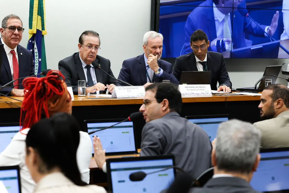 Parlamentares participam de audiência pública na Câmara dos Deputados, com mesa diretora, computadores abertos e bandeira do Brasil ao fundo.