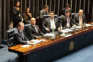 Parlamentares participam de sessão no Senado Federal, sentados à mesa principal com documentos, copos de água e assessores ao fundo.