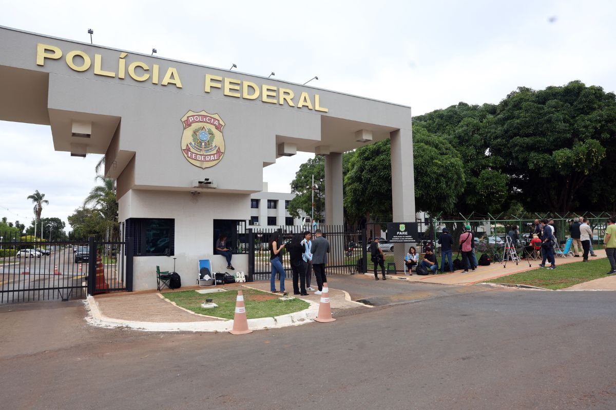 Entrada da sede da Polícia Federal com pessoas reunidas do lado de fora, viaturas próximas e jornalistas acompanhando movimentação.