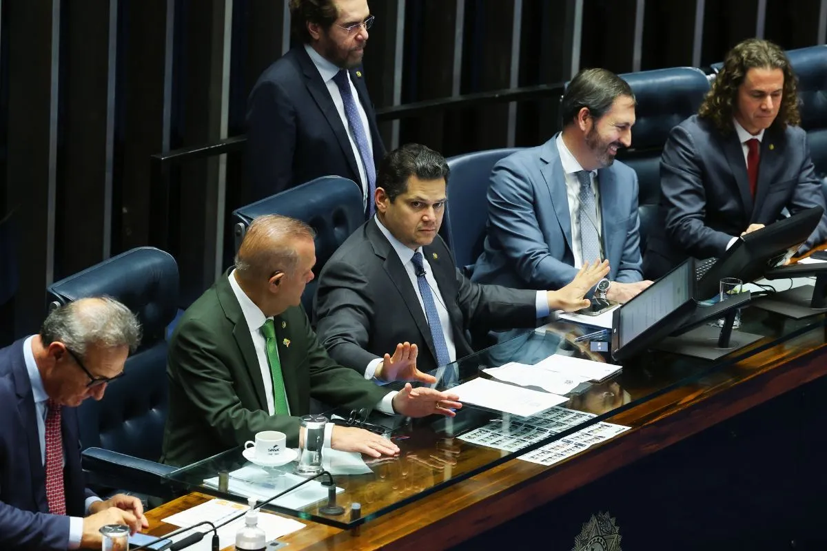 Parlamentares sentados na mesa diretora do Senado Federal durante sessão legislativa, com documentos, painéis eletrônicos e gestos de deliberação.