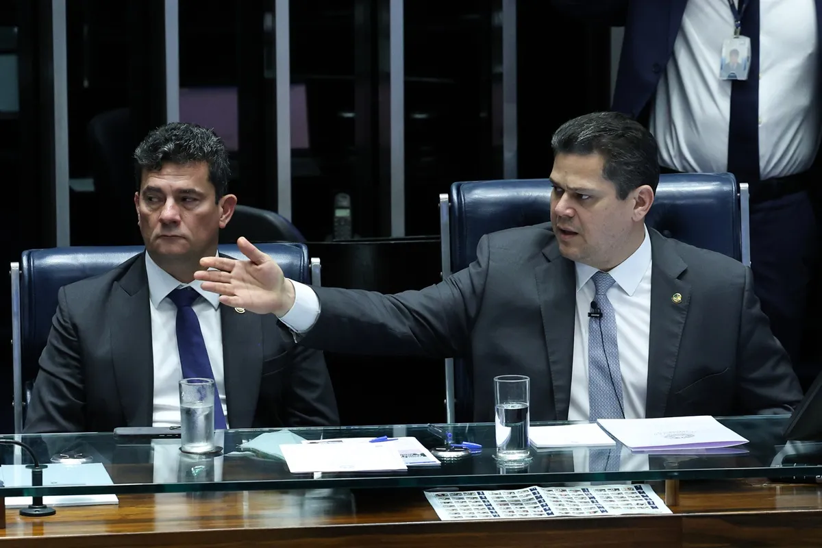Dois senadores sentados no plenário do Senado Federal durante sessão, um deles gesticulando enquanto debate, com copos de água e documentos à mesa.