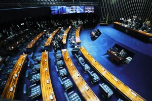 Vista ampla do plenário do Senado Federal em sessão, com mesas de madeira em semicírculo, parlamentares e mesa diretora ao fundo.