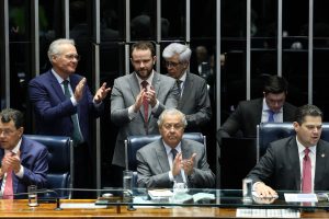 Parlamentares aplaudem durante sessão no plenário do Congresso Nacional, com mesa diretora, documentos e computadores em uso.