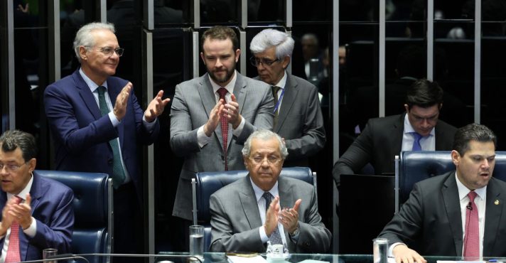 Parlamentares aplaudem durante sessão no plenário do Congresso Nacional, com mesa diretora, documentos e computadores em uso.