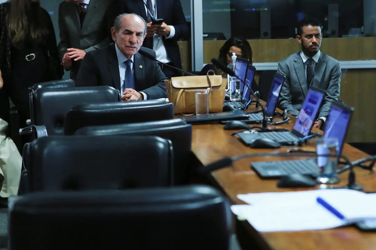Homem sentado em mesa de audiência pública, em ambiente legislativo, com notebooks abertos, microfones e assessores ao redor.