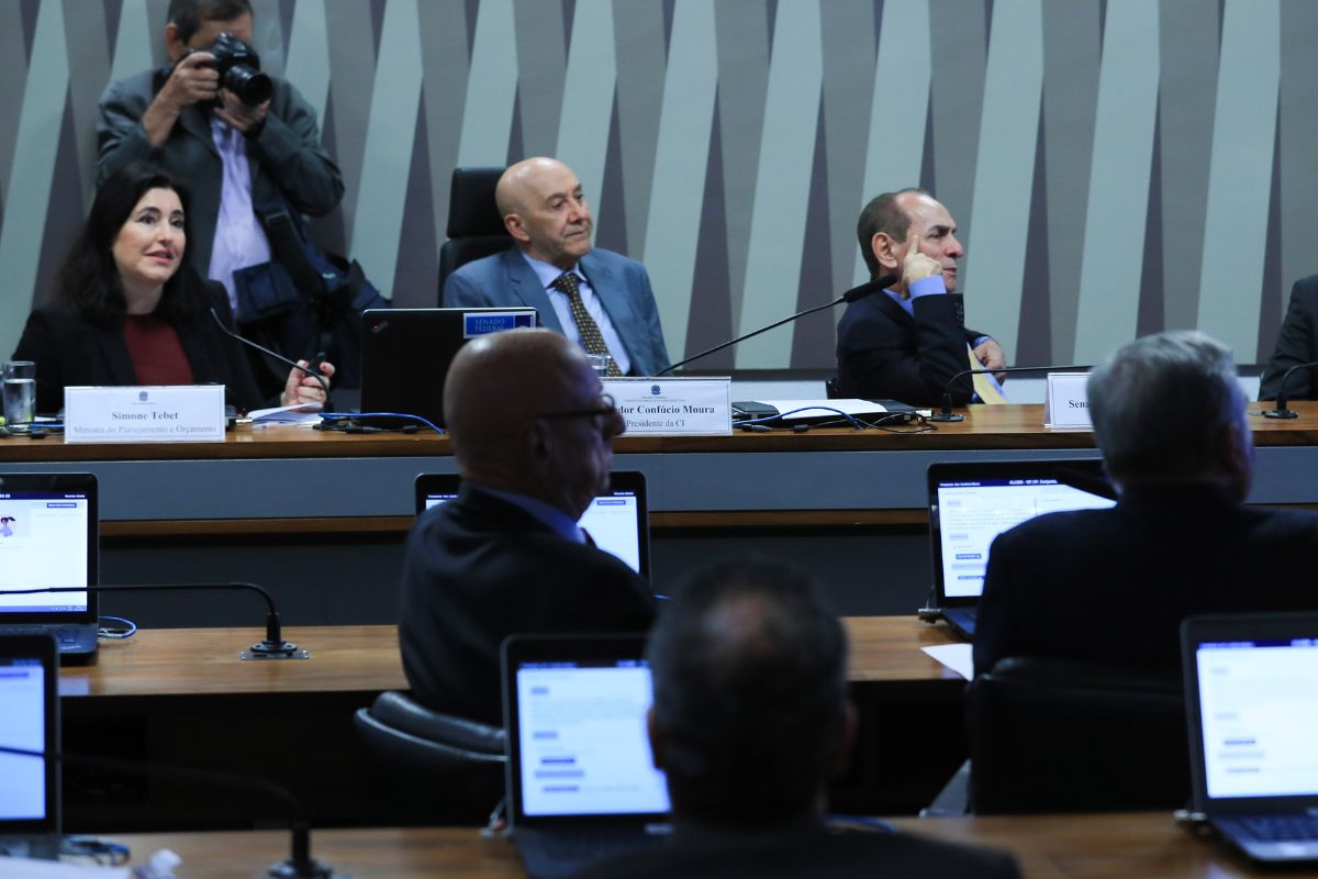 Audiência pública no Senado com autoridades sentadas à mesa, notebooks abertos, microfones e participantes acompanhando o debate sobre orçamento público.