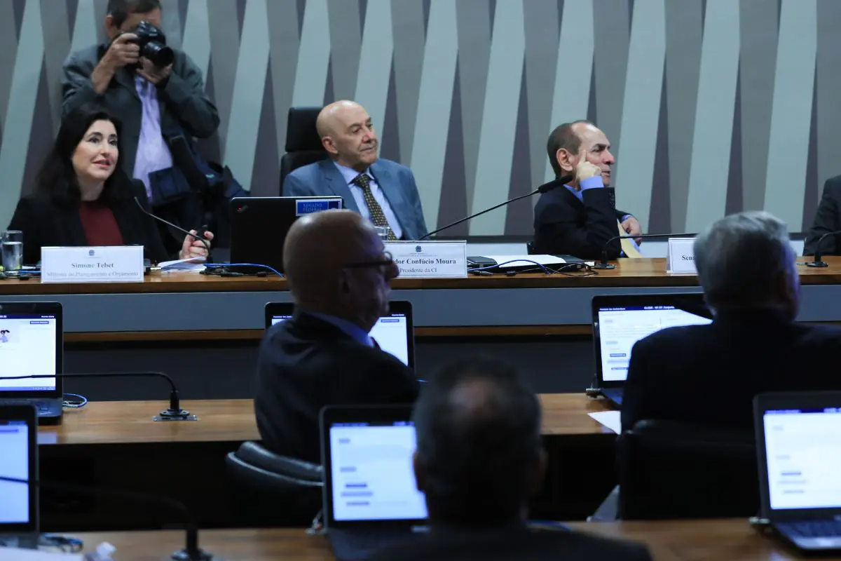 Audiência pública no Senado com autoridades sentadas à mesa, notebooks abertos, microfones e participantes acompanhando o debate sobre orçamento público.