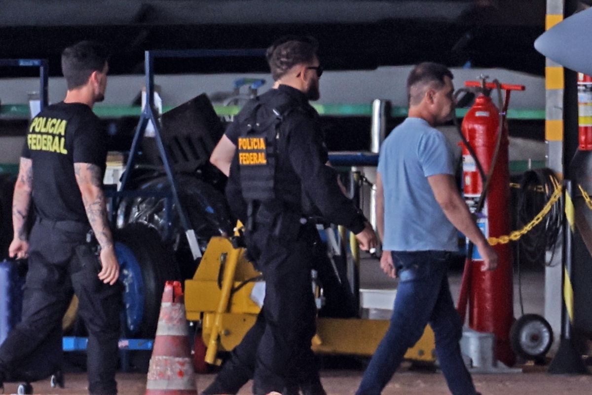 Agentes da Polícia Federal escoltam homem durante operação policial em área industrial, com equipamentos e viaturas ao redor.