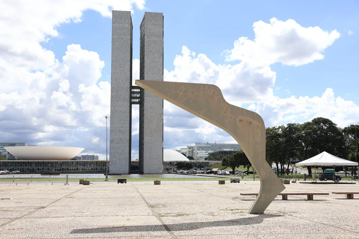 Vista do Congresso Nacional em Brasília com escultura moderna em primeiro plano e prédios institucionais ao fundo.