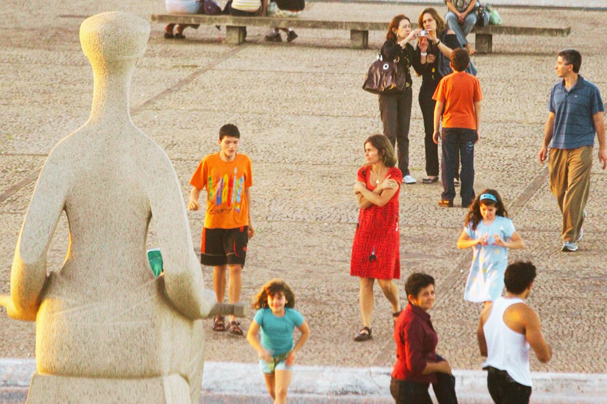 Pessoas circulando na Praça dos Três Poderes em Brasília, com a escultura da Justiça em primeiro plano