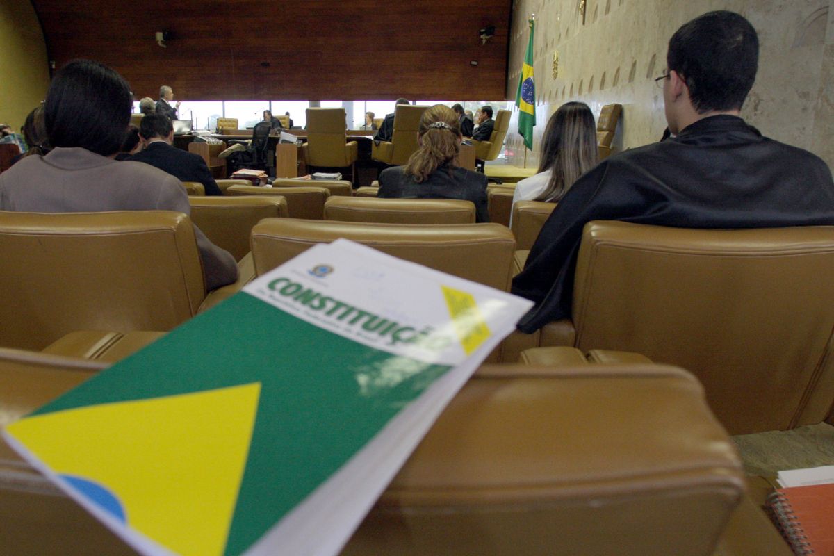 Exemplar da Constituição Federal em primeiro plano durante sessão no Supremo Tribunal Federal, com ministros e público ao fundo.