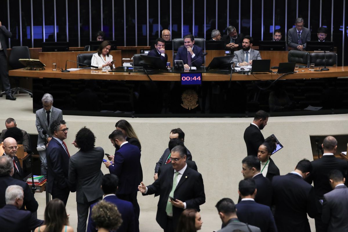 Plenário da Câmara dos Deputados durante sessão sobre impostos no Brasil, com parlamentares reunidos, mesa diretora ao fundo e painel de tempo em destaque.