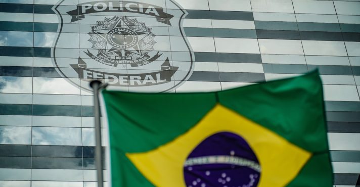 Fachada da Polícia Federal com escudo oficial ao fundo e bandeira do Brasil em primeiro plano, representando atuação institucional.
