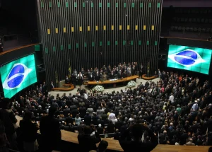 Plenário do Congresso Nacional lotado durante sessão solene, com autoridades reunidas à mesa diretora e bandeira do Brasil exibida em painéis.