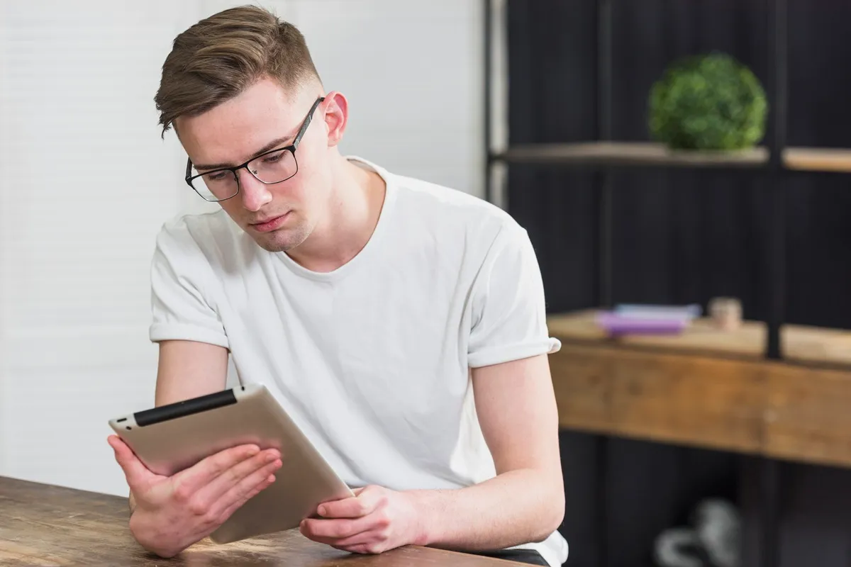Jovem usando óculos lendo informações em tablet enquanto está sentado à mesa em ambiente interno moderno.