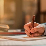 Mão de uma pessoa segurando uma caneta e assinando um documento em uma mesa, representando formalização de contrato ou ato legal