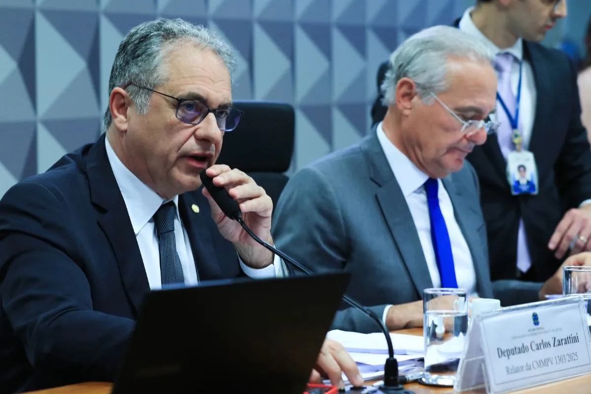 Parlamentar fala ao microfone durante reunião de comissão na Câmara dos Deputados, com documentos, notebook e outro parlamentar ao lado.