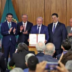 Autoridades do governo federal seguram documento oficial assinado durante cerimônia solene, com bandeiras do Brasil ao fundo e público acompanhando o ato.