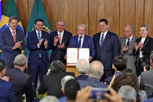 Autoridades do governo federal seguram documento oficial assinado durante cerimônia solene, com bandeiras do Brasil ao fundo e público acompanhando o ato.