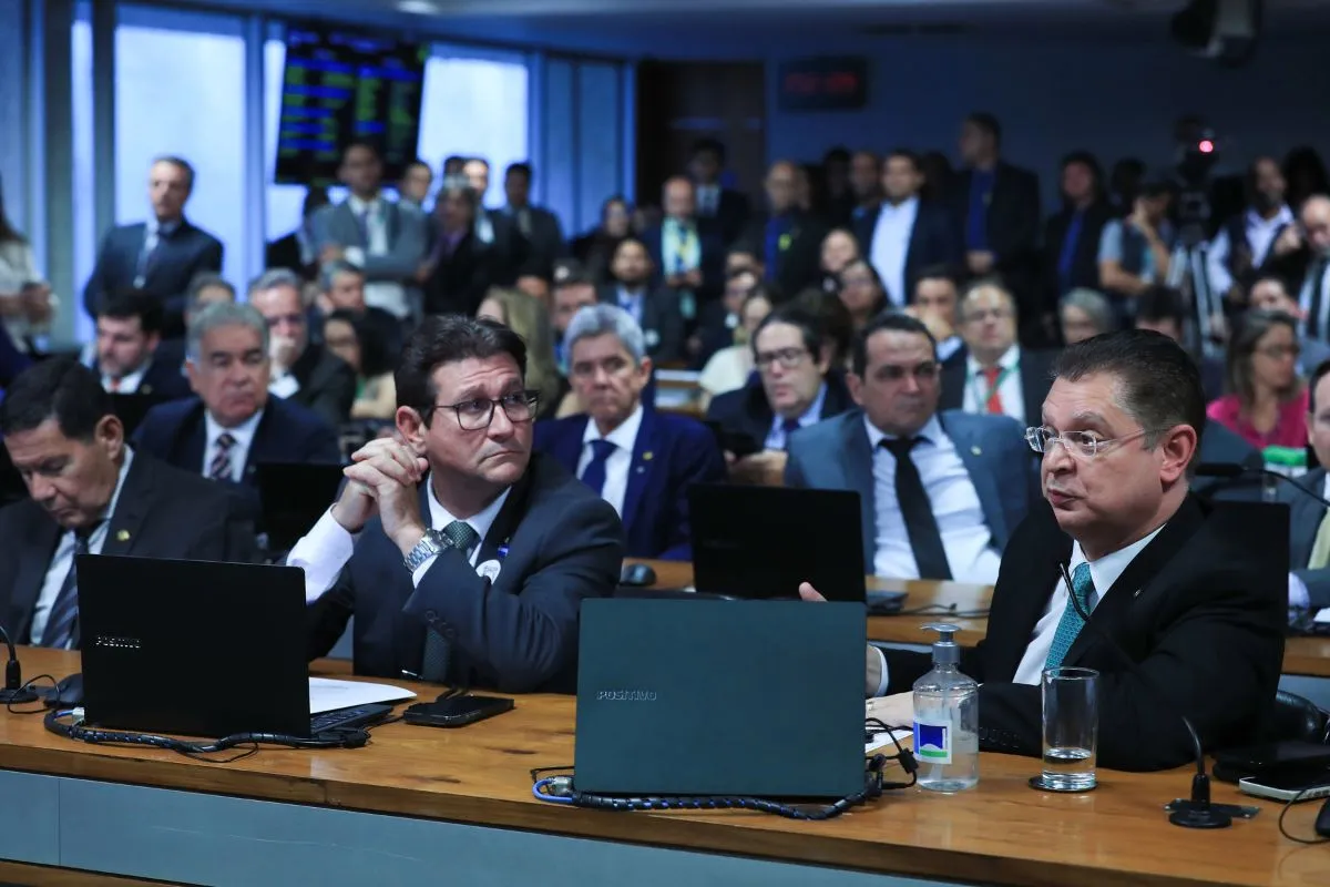 Parlamentares participam de audiência pública em plenário, sentados com notebooks abertos durante debate legislativo, com público ao fundo.
