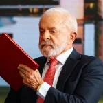 Presidente Luiz Inácio Lula da Silva segura e lê um documento vermelho durante evento oficial, usando terno escuro e gravata vermelha.