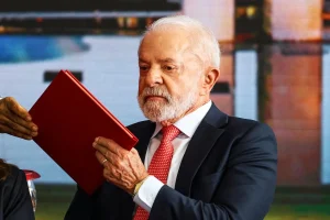 Presidente Luiz Inácio Lula da Silva segura e lê um documento vermelho durante evento oficial, usando terno escuro e gravata vermelha.