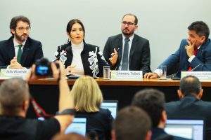 Autoridade feminina fala em audiência pública sobre orçamento público, sentada à mesa com parlamentares, microfones e público acompanhando.
