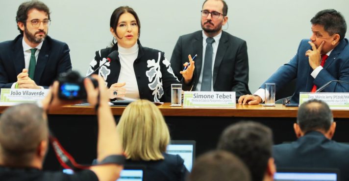 Autoridade feminina fala em audiência pública sobre orçamento público, sentada à mesa com parlamentares, microfones e público acompanhando.