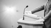 Homem de terno discursando em um púlpito de madeira com um livro aberto, usando microfone em ambiente formal e minimalista.