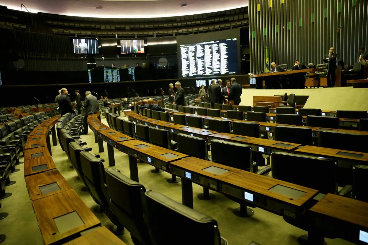 Plenário legislativo com cadeiras e bancadas vazias enquanto parlamentares conversam antes do início da sessão