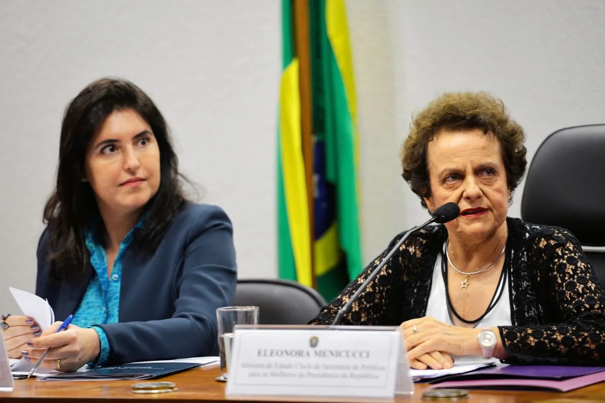 Duas parlamentares sentadas à mesa de comissão da Câmara durante audiência pública, uma delas falando ao microfone, com bandeira do Brasil ao fundo.