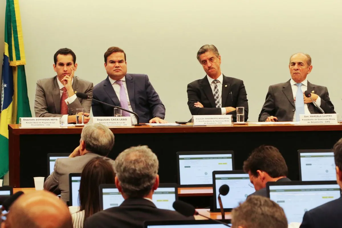 Mesa de comissão legislativa com parlamentares sentados durante sessão oficial, público acompanhando com notebooks e microfones no plenário.