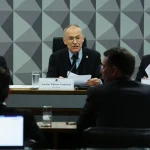 Presidente de comissão parlamentar fala ao microfone durante audiência pública no Senado, com documentos sobre a mesa