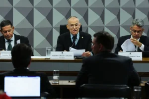 Presidente de comissão parlamentar fala ao microfone durante audiência pública no Senado, com documentos sobre a mesa